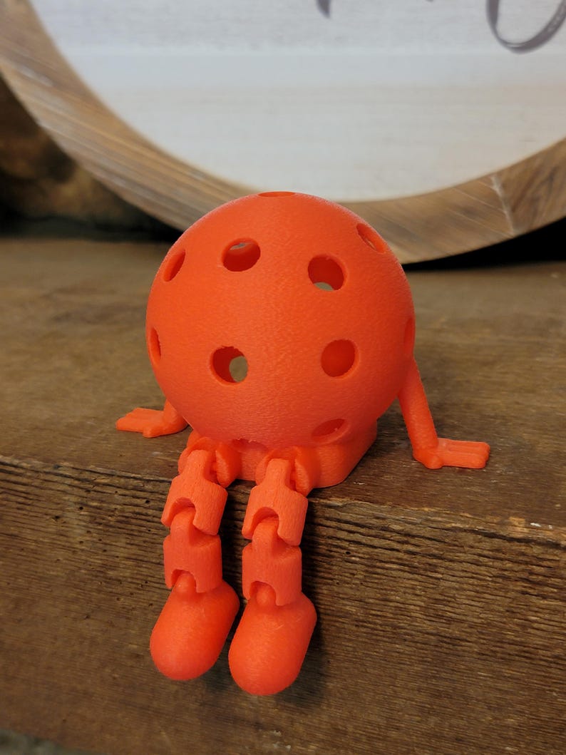 May include: An orange, spherical, 3D-printed figure with a perforated surface, articulated legs, and small arms. The figure is sitting on a wooden surface. The design resembles a whimsical character or toy.