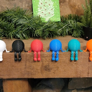 Op de afbeelding: Een rij van zes kleurrijke golfbalfiguren met armen en benen. De golfballen zijn wit, zwart, rood, blauw, turquoise en oranje. Ze staan op een houten oppervlak, met groen en een kerstkaart op de achtergrond.