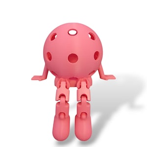 May include: A pink, spherical, articulated toy figure with a perforated head, small arms, and jointed legs. The toy is designed to sit, with its legs bent at the knees. The figure is made of a solid, smooth material and casts a shadow.