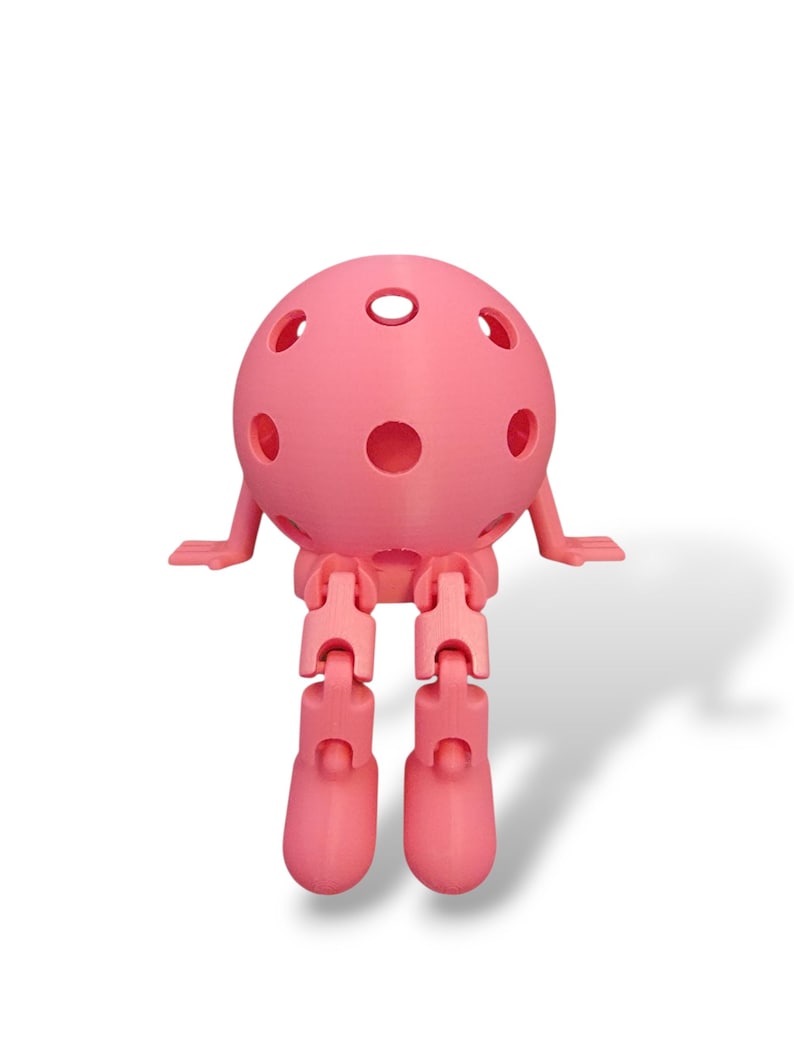 May include: A pink, spherical, articulated toy figure with a perforated head, small arms, and jointed legs. The toy is designed to sit, with its legs bent at the knees. The figure is made of a solid, smooth material and casts a shadow.
