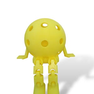 May include: A yellow, spherical toy with multiple holes, small arms, and articulated legs and feet. The toy is designed to sit upright. The toy is made of a solid, matte material and is set against a white background.