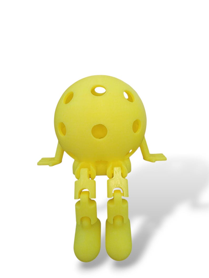 May include: A yellow, spherical toy with multiple holes, small arms, and articulated legs and feet. The toy is designed to sit upright. The toy is made of a solid, matte material and is set against a white background.
