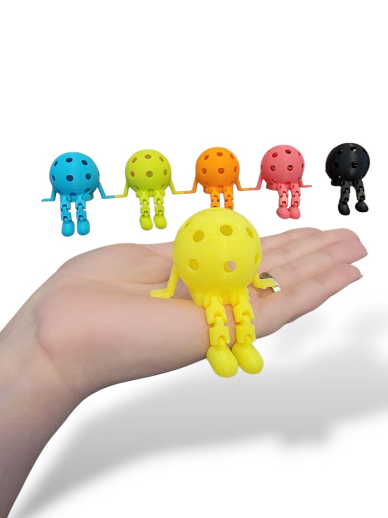 Pu&ograve; includere: Una collezione di figurine colorate stampate in 3D, ciascuna con una testa sferica e gambe articolate. Le figurine sono in vari colori: blu, verde, arancione, rosa, giallo e nero. La figurina gialla &egrave; in primo piano, con le altre disposte dietro.