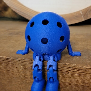 May include: A blue, 3D-printed articulated figure with a spherical head featuring several circular holes. The figure has small, flat feet and arms, and jointed legs. It is sitting on a wooden surface.