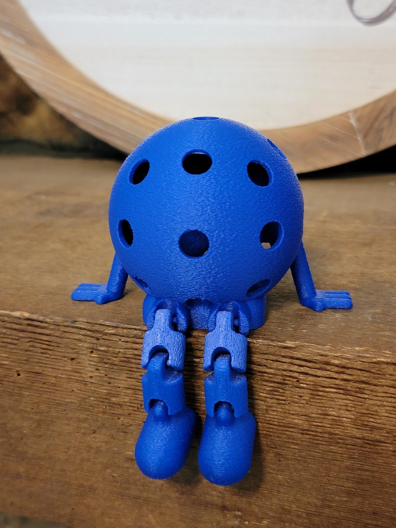 May include: A blue, 3D-printed articulated figure with a spherical head featuring several circular holes. The figure has small, flat feet and arms, and jointed legs. It is sitting on a wooden surface.