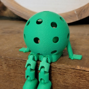 May include: A green, 3D-printed figure resembling a pickleball with articulated legs and arms. The figure has a textured surface and several circular holes. It is sitting on a wooden surface.