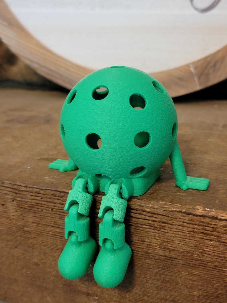 May include: A green, 3D-printed figure resembling a pickleball with articulated legs and arms. The figure has a textured surface and several circular holes. It is sitting on a wooden surface.