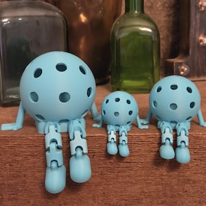 May include: Three light blue spherical figures with circular cutouts, articulated arms, and jointed legs. Each figure features a round head with multiple holes, thin arms extending to the sides, and dangling legs with rounded feet in a seated pose.