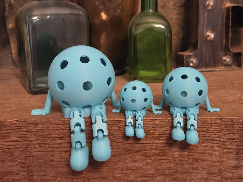 May include: Three light blue spherical figures with circular cutouts, articulated arms, and jointed legs. Each figure features a round head with multiple holes, thin arms extending to the sides, and dangling legs with rounded feet in a seated pose.