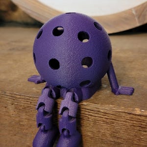 May include: A purple, 3D-printed articulated figure with a spherical head dotted with holes. The figure has jointed legs and arms, and is posed as if sitting. The figure is made of a textured plastic material.