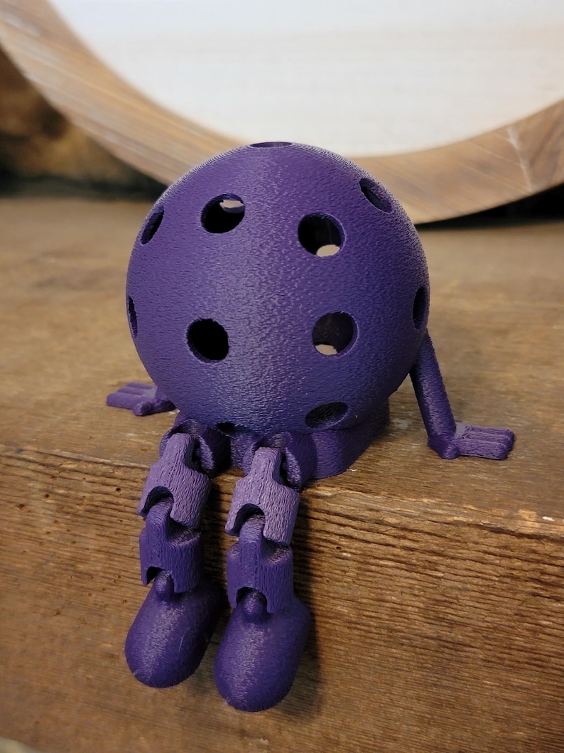 May include: A purple, 3D-printed articulated figure with a spherical head dotted with holes. The figure has jointed legs and arms, and is posed as if sitting. The figure is made of a textured plastic material.