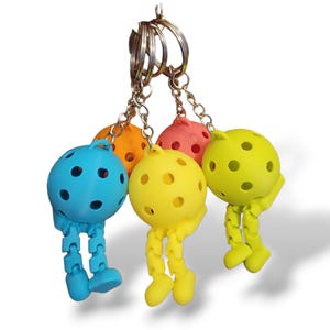 Può includere: Una collezione di portachiavi colorati con figure rotonde, perforate a forma di palla con gambe e braccia articolate. I portachiavi sono blu, arancioni, rosa, gialli e verdi, attaccati ad anelli portachiavi argentati.