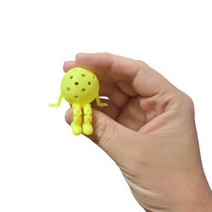 Pu&ograve; includere: Un piccolo giocattolo giallo a forma di palla con fori multipli, braccia e gambe articolate. Il giocattolo &egrave; tenuto in mano su sfondo bianco. Il design del giocattolo assomiglia a un personaggio dei cartoni animati.