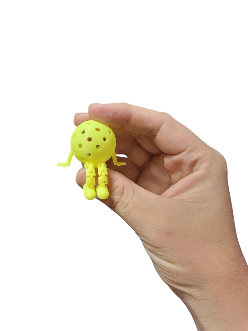 Pu&ograve; includere: Un piccolo giocattolo giallo a forma di palla con fori multipli, braccia e gambe articolate. Il giocattolo &egrave; tenuto in mano su sfondo bianco. Il design del giocattolo assomiglia a un personaggio dei cartoni animati.