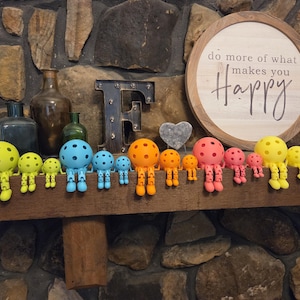 May include: A collection of articulated figures with spherical heads resembling pickleballs, arranged in a row by color including lime green, blue, orange, pink, and yellow. Each figure features jointed limbs and sits on a ledge in front of a sign.