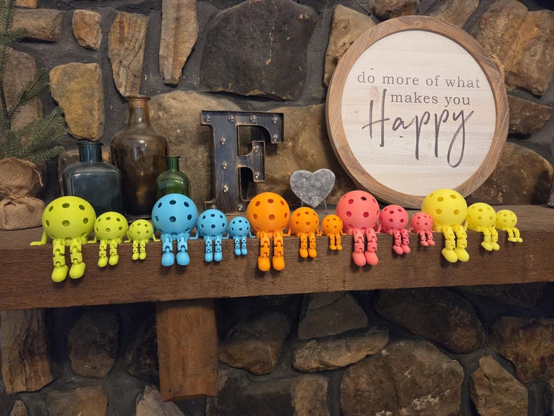 May include: A collection of articulated figures with spherical heads resembling pickleballs, arranged in a row by color including lime green, blue, orange, pink, and yellow. Each figure features jointed limbs and sits on a ledge in front of a sign.