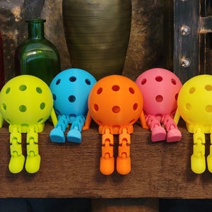 May include: A collection of colorful, spherical toys with perforated surfaces and articulated legs. The toys are in shades of yellow, blue, orange, and pink, and are arranged on a wooden surface. The toys have a whimsical, playful design.