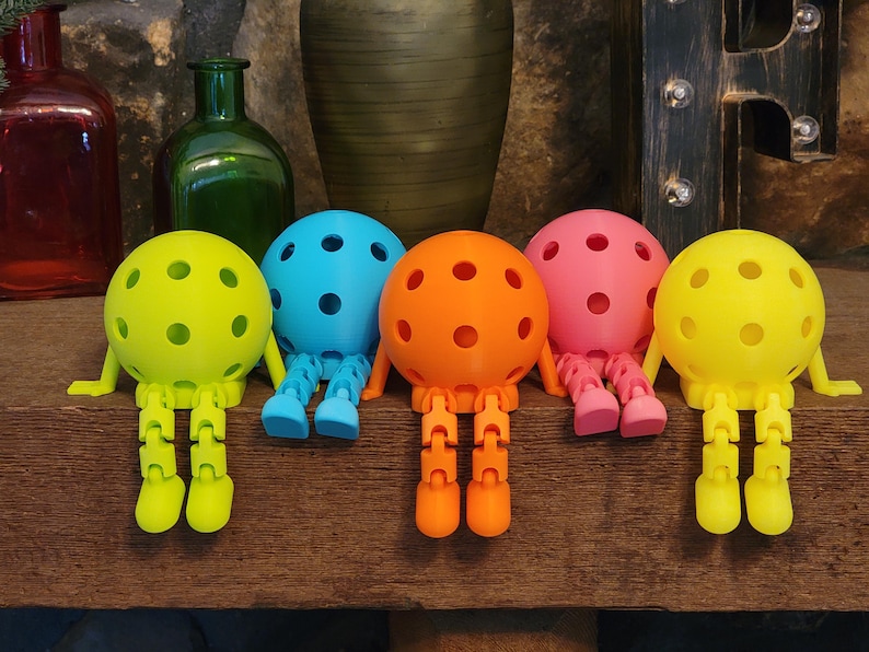 May include: A collection of colorful, spherical toys with perforated surfaces and articulated legs. The toys are in shades of yellow, blue, orange, and pink, and are arranged on a wooden surface. The toys have a whimsical, playful design.