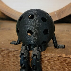 May include: A black, spherical decorative object with multiple circular holes, resembling a ball. It has small arms and articulated legs, giving it a whimsical, seated appearance. The object is placed on a wooden surface.
