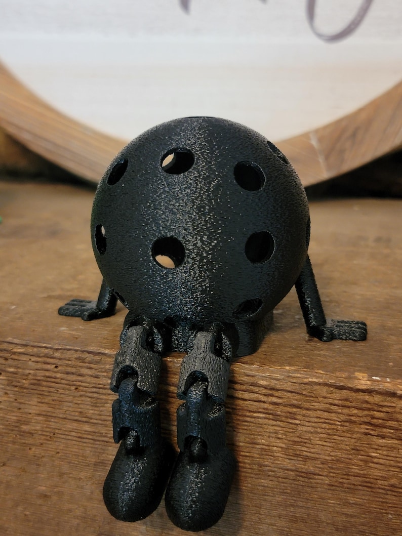 May include: A black, spherical decorative object with multiple circular holes, resembling a ball. It has small arms and articulated legs, giving it a whimsical, seated appearance. The object is placed on a wooden surface.