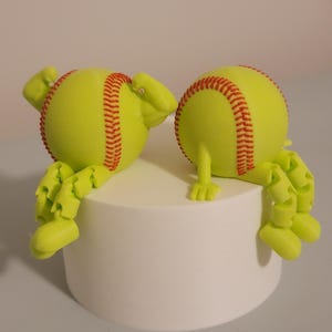 May include: Two lime green softball-shaped figures with red stitching details. The figures have articulated limbs and are positioned on a white cylindrical base. The figures appear to be interacting with each other.
