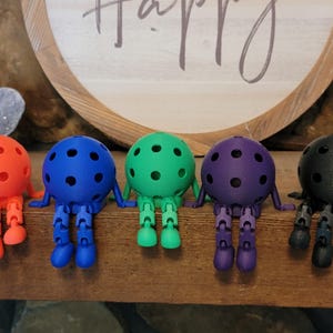 May include: Five colorful, articulated ball-shaped figures with legs and arms are arranged on a wooden surface. The figures are in shades of orange, blue, green, purple, and black. Each has a series of small, dark dots on its spherical body.