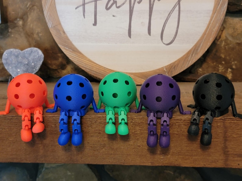 May include: Five colorful, articulated ball-shaped figures with legs and arms are arranged on a wooden surface. The figures are in shades of orange, blue, green, purple, and black. Each has a series of small, dark dots on its spherical body.