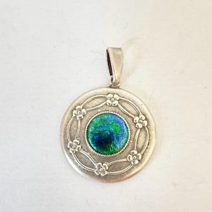 Puede incluir: Colgante redondo de plata con una piedra central verde y azul iridiscente. El colgante está rodeado por un diseño floral y tiene un lazo para una cadena. Probablemente de plata de ley.