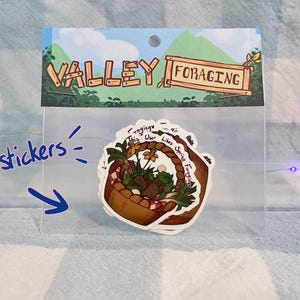 May include: A clear plastic package containing four stickers. The stickers feature a brown basket filled with plants and flowers. The package has a header with the words "Valley Foraging".
