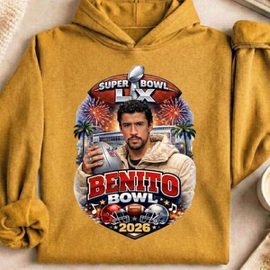 May include: Mustard yellow hoodie featuring a graphic of a man holding a football, with the text "Benito Bowl 2026" and "Super Bowl LX". The design includes football helmets, fireworks, and palm trees.