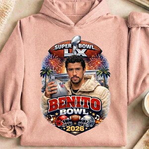 May include: A light pink hoodie featuring a graphic of a man holding a football, with the text "BENITO BOWL 2026". The design includes a football, helmets, fireworks, and palm trees, with "SUPER BOWL LX" at the top.
