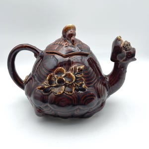 May include: A brown ceramic teapot with a floral design and a unique spout. The teapot has a rounded body, a curved handle, and a lid with a small figure on top. The teapot is a dark brown color with lighter brown accents.
