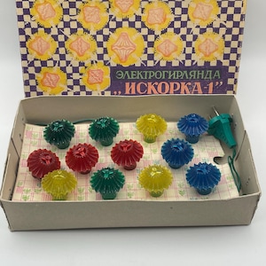 May include: A vintage string of Christmas lights in a box. The lights are various colours, including red, green, yellow, and blue. The box has a purple and white chequered pattern with yellow floral designs. Text on the box reads "ЭЛЕКТРОГИРЛЯНДА, ИСКОРКА-1".
