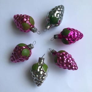 May include: Six vintage Christmas ornaments shaped like grapes. The ornaments are a mix of shiny silver and bright pink with green leaf accents. Each ornament has a small metal loop for hanging.