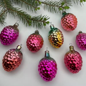 May include: A collection of eight glass pinecone ornaments in shades of pink, magenta, and gold with a textured, bumpy surface. Each ornament features a silver-toned metal cap at the top for hanging, arranged below a green pine tree branch.