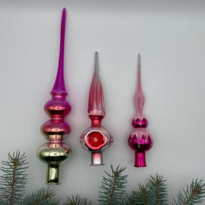 Può includere: Tre puntali per albero in vetro disposti verticalmente in varie tonalità di rosa e argento. Il puntale più alto presenta una lunga guglia con una sfumatura dal rosa scuro all'argento metallizzato. I due più piccoli hanno basi bulbose.