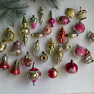 Può includere: Una collezione di decorazioni natalizie vintage di varie forme e colori, tra cui rosa, oro e argento. Gli ornamenti sono disposti su una superficie bianca, con alcuni rametti di sempreverdi nell'angolo in alto a sinistra.