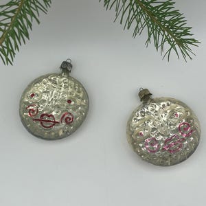 May include: Two round, silver-toned glass ornaments featuring embossed sun-like faces with painted red details. Each ornament has a metallic cap at the top for hanging, with a textured, reflective surface that catches light, creating a vintage appearance.