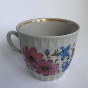 Op de afbeelding: Een witte keramische theekop met een gouden rand en een oor. De kop is versierd met roze en blauwe bloemmotieven. De kop heeft een geribbelde textuur aan de buitenkant.