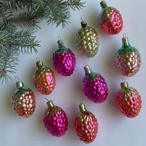 May include: A collection of vintage Christmas ornaments shaped like pine cones. The ornaments are in various colors, including pink, gold, and green, with a metallic finish. A sprig of evergreen branches is in the upper left corner.
