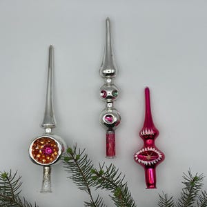 May include: Three vintage glass tree toppers. One silver topper features a large round centre with orange and pink textured details. A second silver topper has stacked spheres with pink accents. A third topper is bright pink with white painted details.