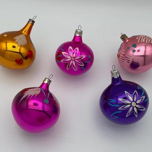 May include: Five glass Christmas ornaments in various shapes and colors. One is orange, two are magenta, one is light pink, and one is purple. Each ornament features hand-painted floral or abstract white and teal designs and a silver metal cap.