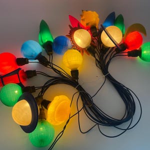 May include: A string of vintage Christmas lights with various shapes and colours. The lights include red rocket, green globe, yellow sun, and blue globe designs. The bulbs are connected by black wires.