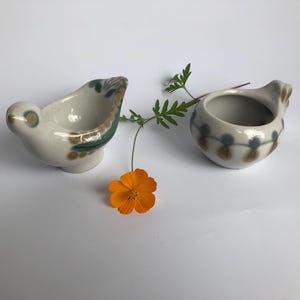 May include: Two ceramic bird-shaped containers with a cream-colored base and decorative patterns in green, brown, and blue. An orange flower and green sprig are placed between the containers. The containers are likely for decorative purposes.