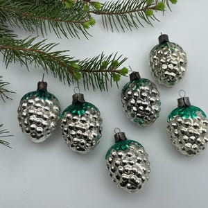 May include: A set of five vintage-style glass ornaments shaped like pinecones. Each ornament features a metallic silver textured body with a dark green cap and a small metal loop at the top for hanging, arranged near a green pine branch.