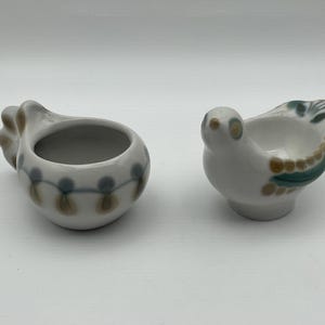 May include: Two ceramic bird-shaped containers. One is a white bird with green and brown accents. The other is a white container with a bulbous shape and a series of brown and blue dots. Both are on a white background.