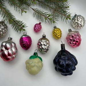 Op de afbeelding: Een verzameling vintage kerstversieringen in verschillende vormen en kleuren, waaronder zilver, roze, geel en blauw. De ornamenten zijn gerangschikt op een wit oppervlak met een takje groenblijvende plant. De ornamenten hebben een textuur, bessenachtig ontwerp.