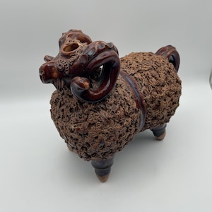 May include: A brown ceramic ram figurine with detailed textured wool and curved horns. The ram has a detailed face and is supported by three legs. The figurine is a decorative item.