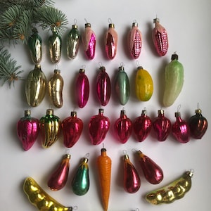 以下が含まれることがあります： 様々な果物や野菜の形をした、ビンテージのガラス製のクリスマスオーナメントのコレクション。オーナメントは緑、赤、黄、ピンクの色合いです。