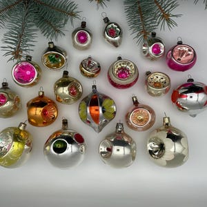 May include: A collection of vintage Christmas ornaments in various shapes and colors, including silver, gold, pink, and orange. Some ornaments have floral or geometric designs. The ornaments are arranged on a white surface with evergreen branches.